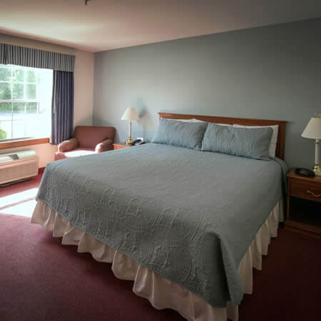 White and light blue painted room with rose carpet, large window, large bed with blue bedspread and an upholstered chair
